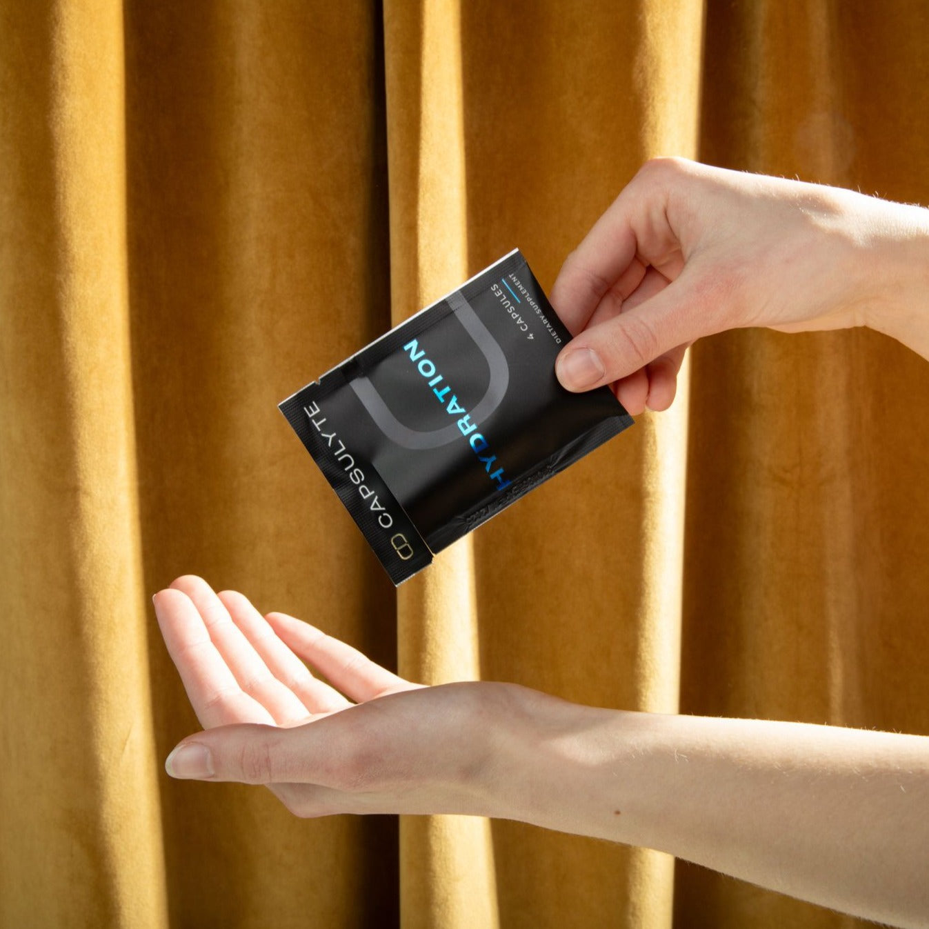 Person pouring Capsulyte Hydration dietary supplement capsules into their hand from a black sachet, against a warm yellow curtain background. The supplement supports hydration and helps prevent hangovers.