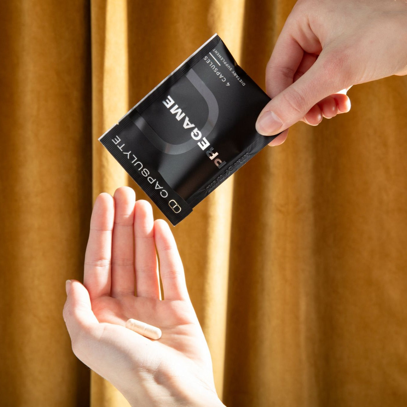 Person pouring Capsulyte Pregame dietary supplement capsules into their hand from a black sachet, set against a warm yellow curtain background. Pregame is designed to prevent hangover symptoms and support the body’s natural defenses after drinking, with key ingredients like DHM, NAC, Siliphos®, and Clovinol®.